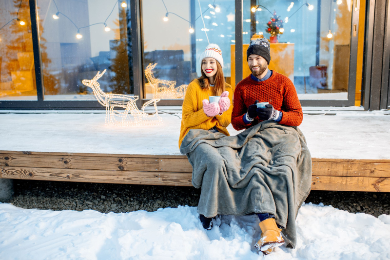 Home 128 A smiling couple in winter clothes sits on a snowy porch, wrapped in blankets and holding mugs. Festive decorations and string lights are behind them, along with a lit reindeer figure.
