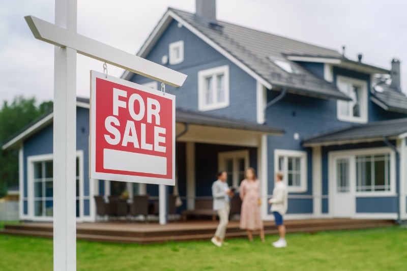 Home 126 A red and white For Sale sign is in the foreground, with a modern blue house and three people talking on the front lawn in the background.