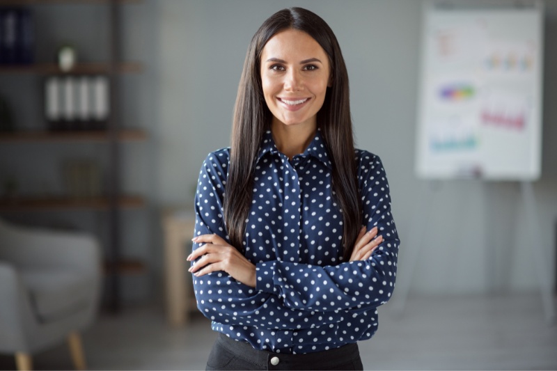 Home 119 A woman with long dark hair, wearing a navy blue polka dot shirt and black pants, stands confidently with arms crossed and smiles in an office setting. A blurred flipchart and shelves are visible in the background.