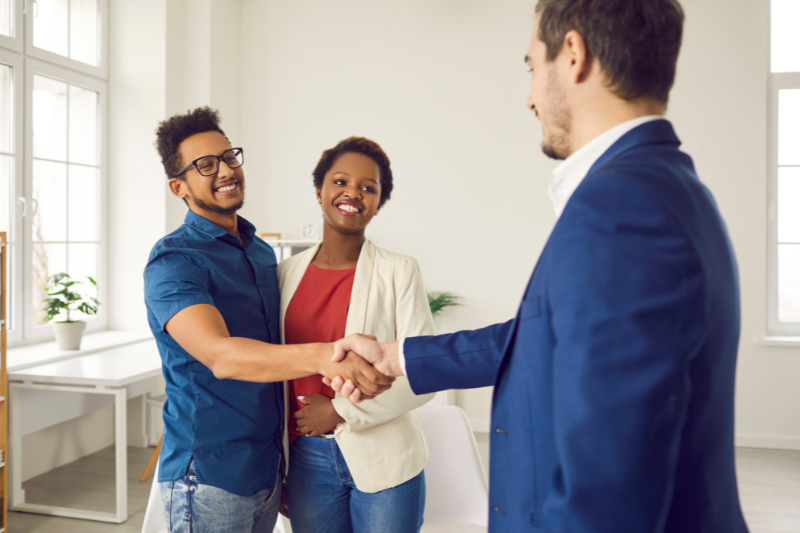 Home 115 A smiling man in a blue shirt shakes hands with a man in a suit while a woman in a white blazer stands beside them, all in a bright office setting.