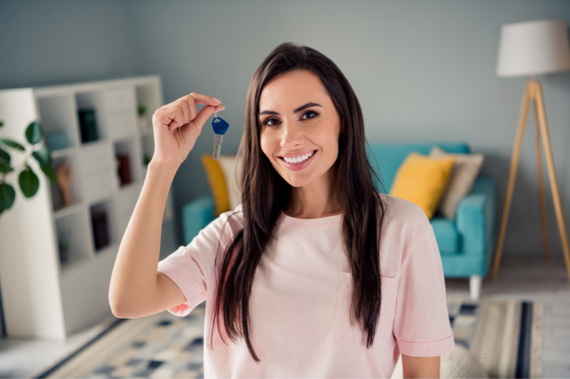 Home 110 A smiling woman with long dark hair holds up a key indoors, standing in a stylish living room with a blue sofa, yellow pillows, a bookshelf, and a floor lamp in the background.