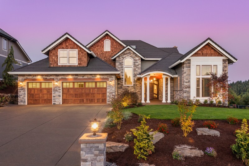 Home 112 A large, modern suburban house with stone and wood exterior, three-car garage with wooden doors, a well-manicured lawn, landscaped garden, and a driveway at sunset.