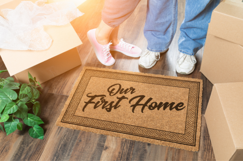 Home 109 Two people stand next to a doormat that reads Our First Home, surrounded by moving boxes and a plant on a wooden floor, with sunlight shining in.
