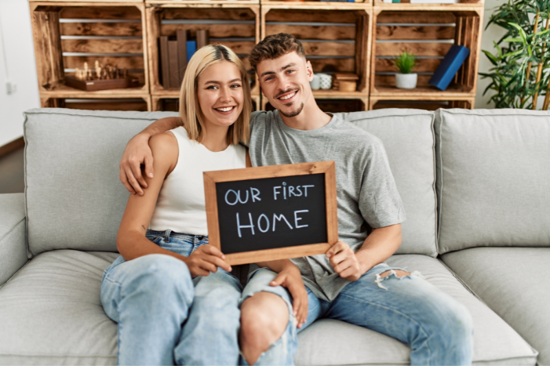 Home 102 A smiling couple sits on a couch holding a chalkboard sign that reads “Our First Home,” with wooden shelves and plants in the background.