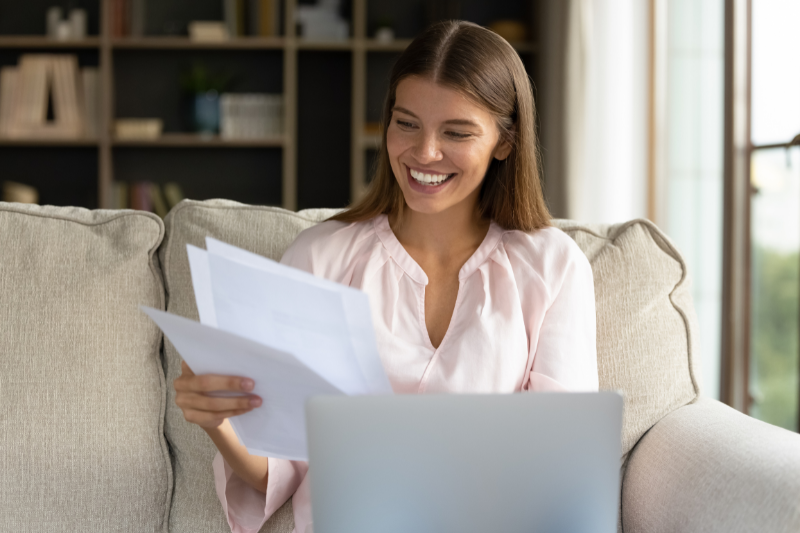 Home 103 A smiling woman sits on a couch holding papers in one hand while looking at a laptop, appearing happy and relaxed in a cozy, well-lit living room with bookshelves in the background.