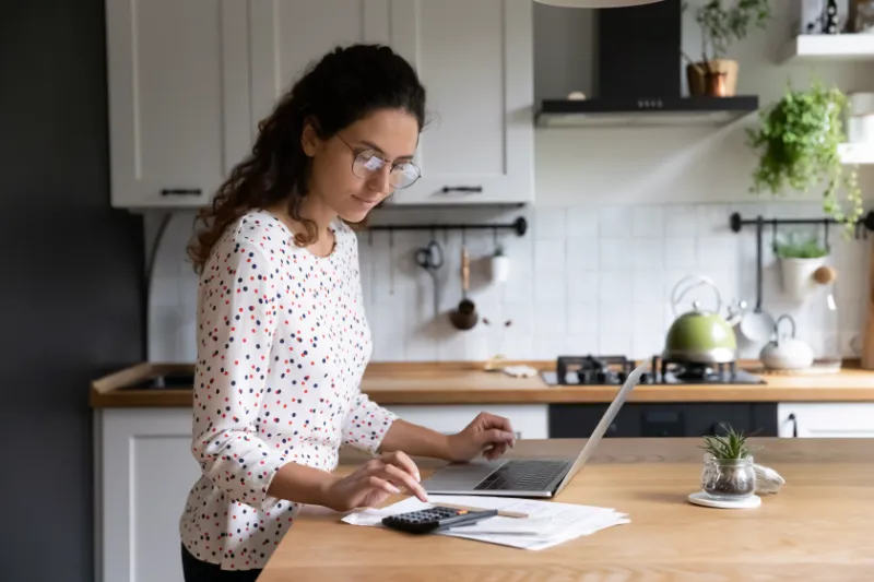 Home 97 A woman stands in a kitchen, using a laptop and a calculator, with papers spread out on the counter. She appears focused, wearing glasses and a polka-dot shirt. Plants and kitchenware are visible in the background.