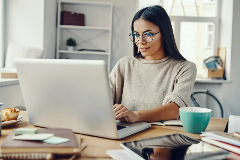 Home 87 A woman with long dark hair and glasses is sitting at a desk, working on a laptop. She is surrounded by notebooks, a mug, and office supplies in a bright, modern workspace.