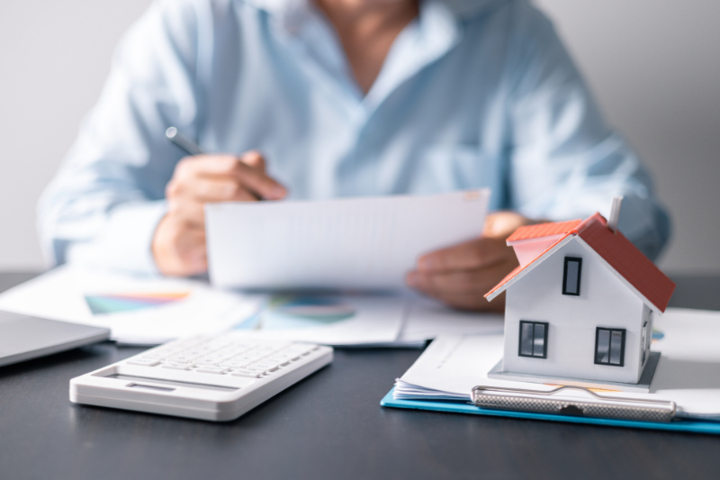 Home 83 A person sits at a desk reviewing documents with charts, next to a calculator and a model house, suggesting work related to real estate or financial planning.