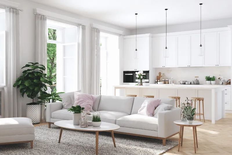 Home 80 A bright, modern open-plan living room and kitchen with white furniture, light wood accents, potted plants, large windows, and pendant lights. Sunlight streams in, creating an airy, inviting atmosphere.