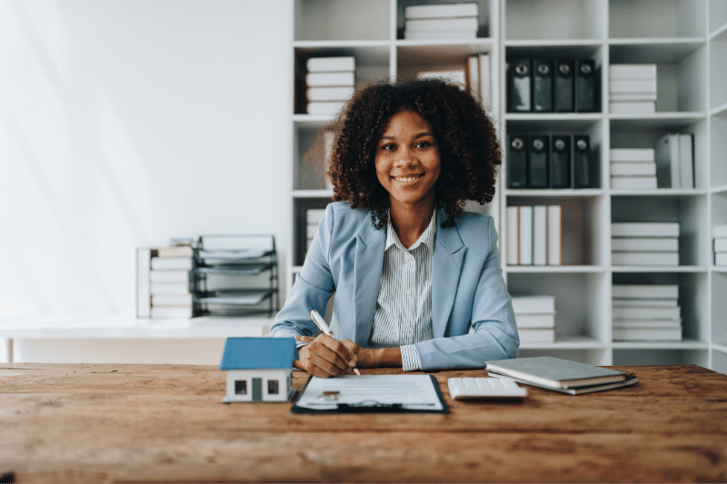 Home 76 A woman in a light blue blazer sits at a desk, smiling, with documents, a pen, a small house model, a notebook, and a calculator in front of her. Bookshelves with files and books are in the background.