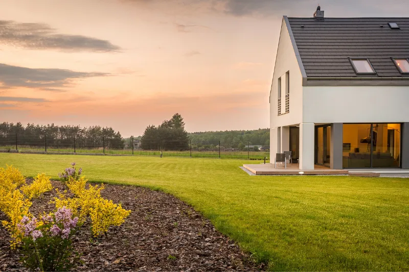 Home 65 Modern house with large windows and sloped roof, set on a spacious green lawn with yellow and purple flowers, under a cloudy sunset sky, with trees and a fence in the background.