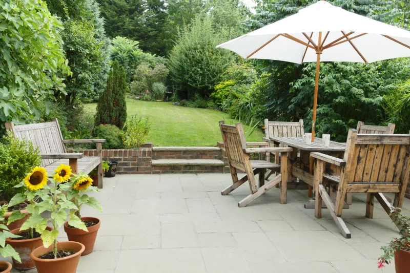 Home 62 A patio with wooden benches and a dining table set beneath a large white umbrella, surrounded by potted sunflowers and lush green garden foliage in the background.