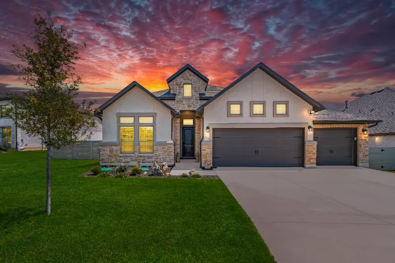 Home 54 A modern, single-story house with stone accents and a three-car garage is shown at sunset. The sky is vibrant with red and purple clouds, and the front lawn is neatly landscaped with a small tree and flower beds.