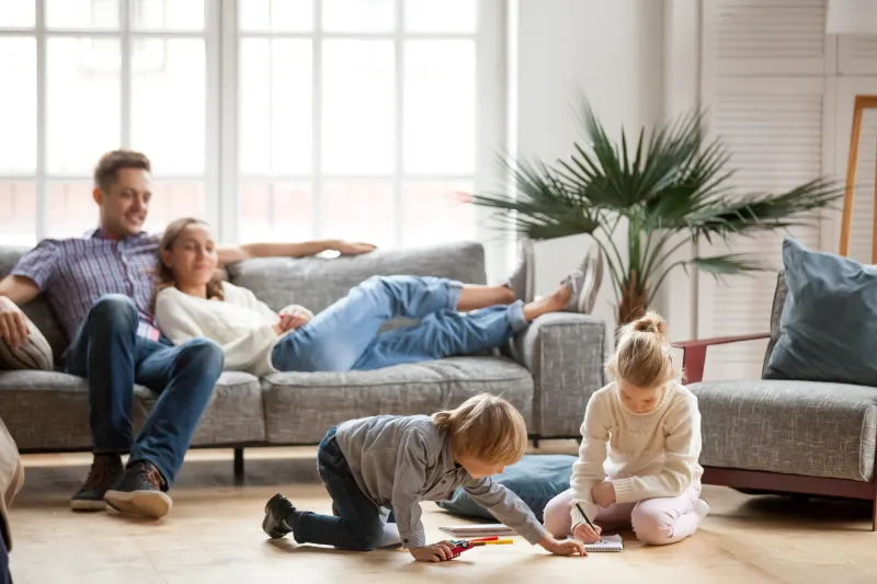 Home 53 A man and woman relax together on a sofa while two young children draw and color on the floor in a bright, cozy living room with large windows and a potted plant.