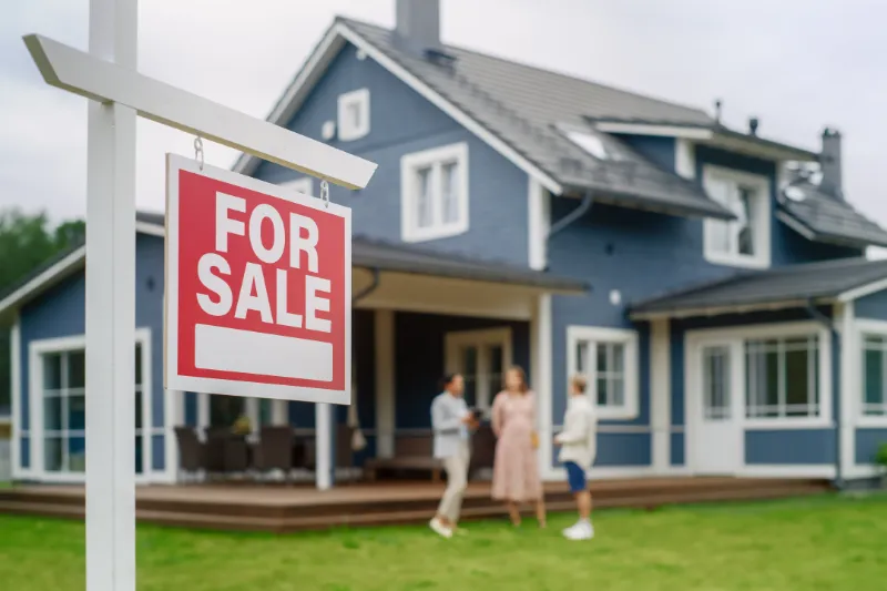 Home 48 A red For Sale sign is displayed in front of a modern blue house. Three people are standing and talking on the porch in the background. The scene suggests a house showing or real estate transaction.