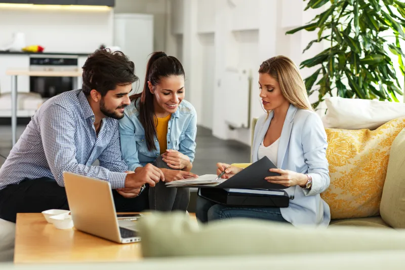 Home 50 A professional woman shows documents to a smiling couple seated on a sofa in a modern, bright living room. A laptop and coffee cup are on the table, and a large plant is in the background.