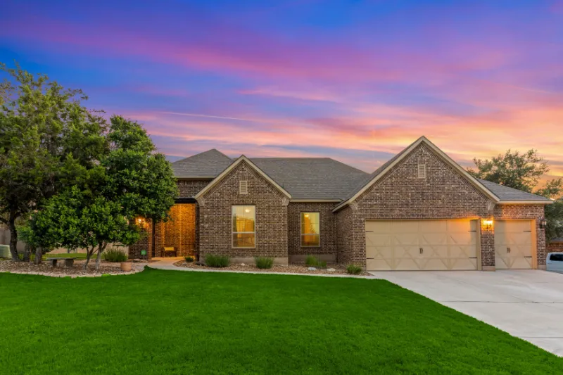 Home 51 Single-story brick house with a three-car garage, lit windows, and a well-kept green lawn, set against a colorful sunset sky with pink and purple hues. Trees and shrubs line the front walkway.