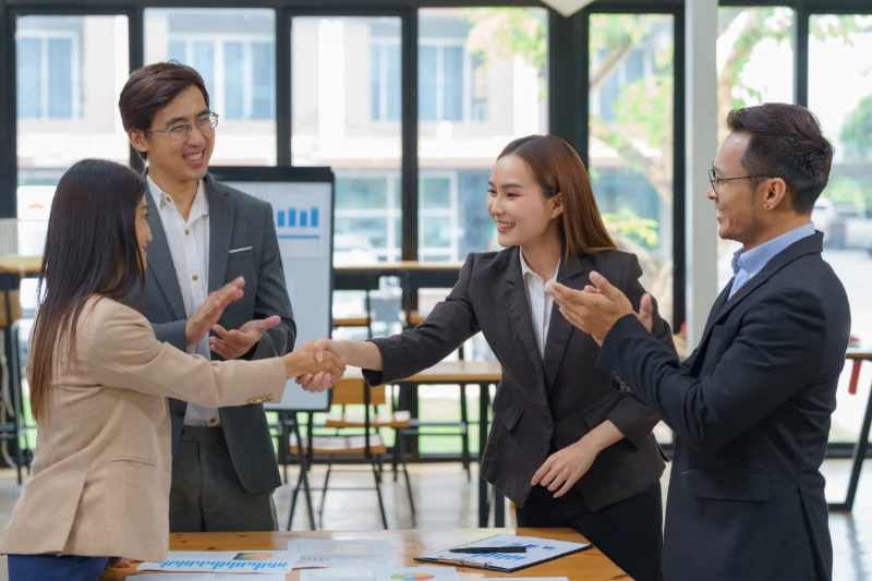 Home 43 Four business professionals in formal attire are in a bright office. Two women in the center are shaking hands, while two men beside them are smiling and clapping, suggesting a successful meeting or agreement.