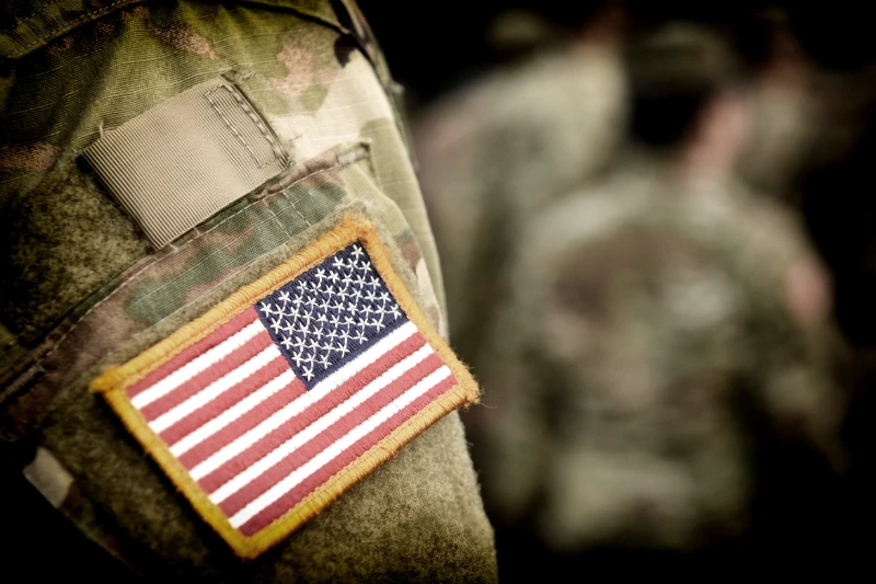 Home 34 Close-up of a U.S. flag patch on the sleeve of a camouflage military uniform, with other blurred soldiers in similar uniforms in the background.