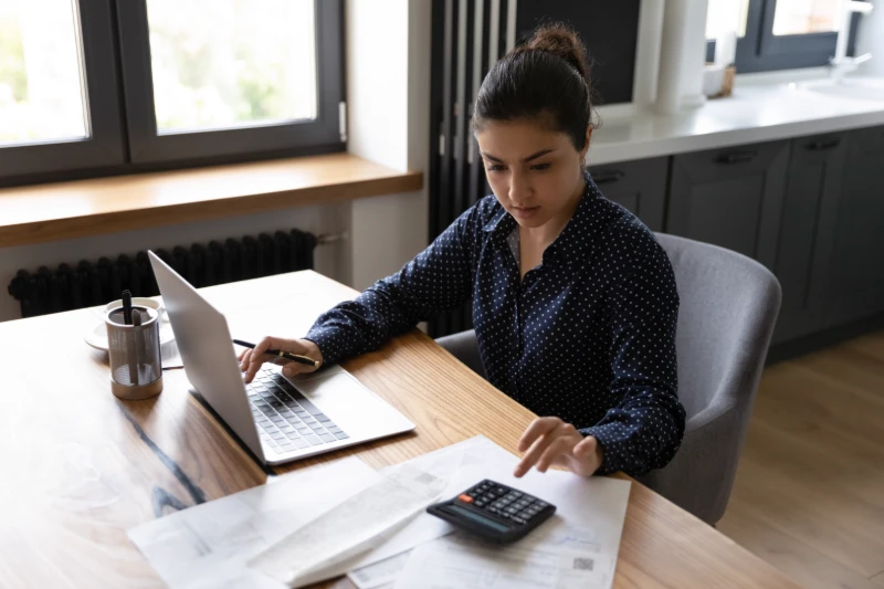Home 27 A woman sits at a table using a laptop and calculator, surrounded by papers and a pen holder, appearing focused while working in a modern home setting with large windows.