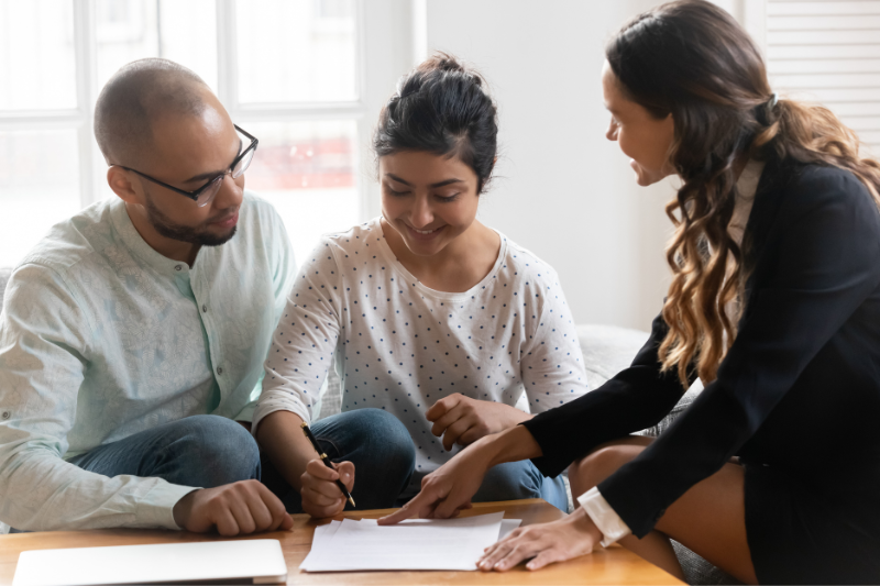 Home 2 A woman in business attire points to a document on a table while a seated man and woman listen and review the paper, appearing to discuss or sign paperwork together in a bright room.