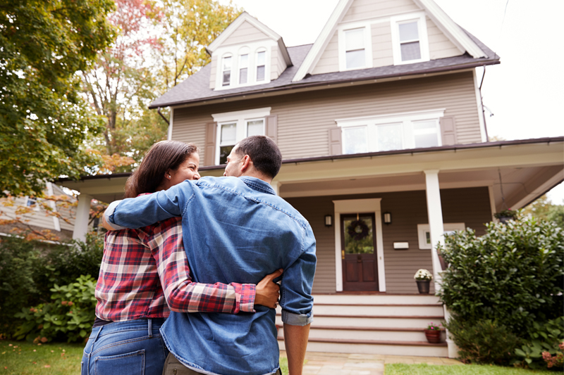 A couple stands outside, arm in arm, looking at a large, two-story house with a porch and green landscaping on a sunny day.