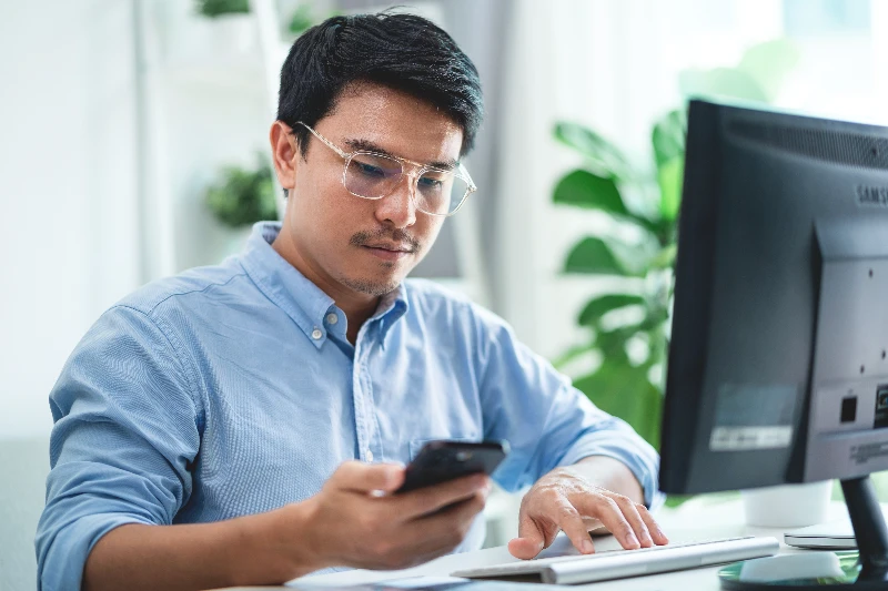 Blog 9 A-man-wearing-glasses-is-sitting-at-a-desk-with-a-computer-monitor-and-a-cell-phone-He-is-looking-at-the-cell-phone-while-typing-on-the-computer