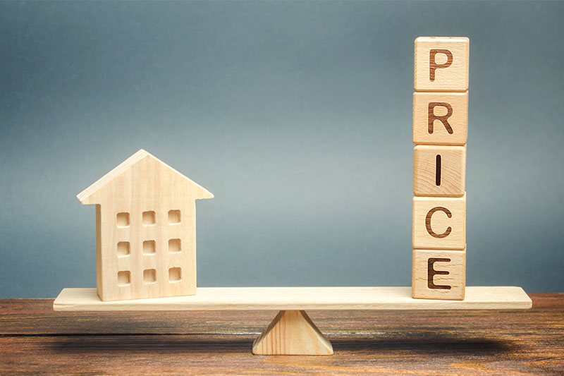 A wooden house model and stacked blocks spelling PRICE are balanced on a seesaw, symbolizing the relationship between property value and price.