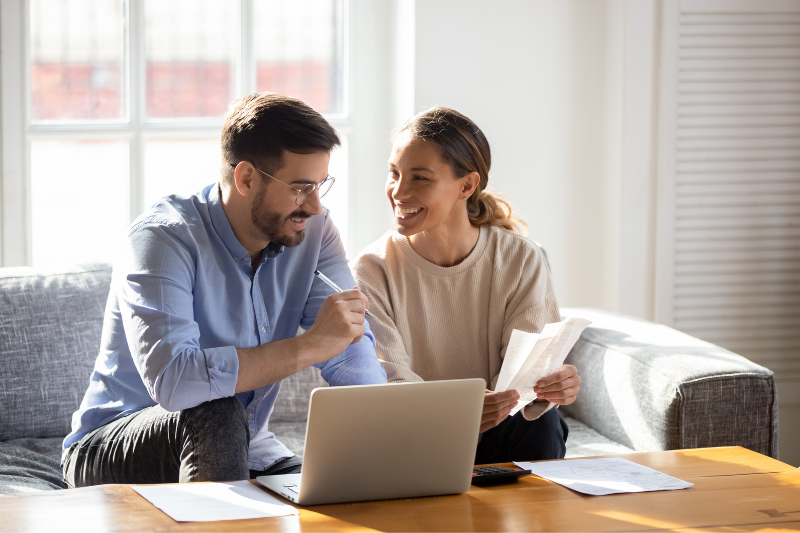 A smiling couple sits together on a couch, looking at documents and a laptop on the coffee table, appearing happy and engaged while discussing paperwork in a bright, sunlit living room.