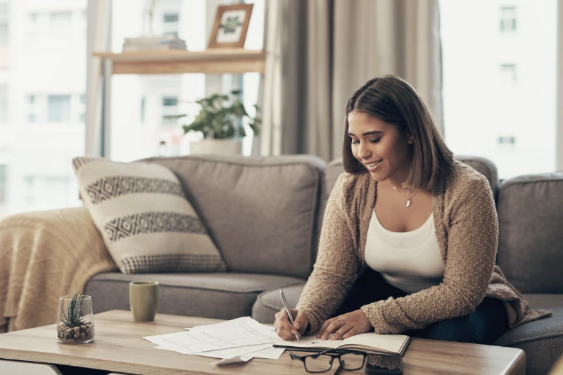 A woman sits on a sofa in a cozy living room, smiling as she writes in a notebook on a coffee table. Papers, a mug, and glasses are also on the table. Shelves and windows are visible in the background.