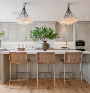 Home 1 Modern kitchen with a large island, three woven bar stools, a vase of green branches as a centerpiece, pendant lights above, and light gray cabinets in the background.